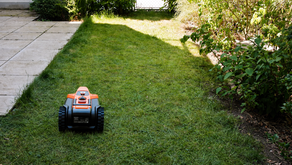 Oranger Mähroboter auf einer schmalen Rasenfläche zwischen Weg und Sträuchern